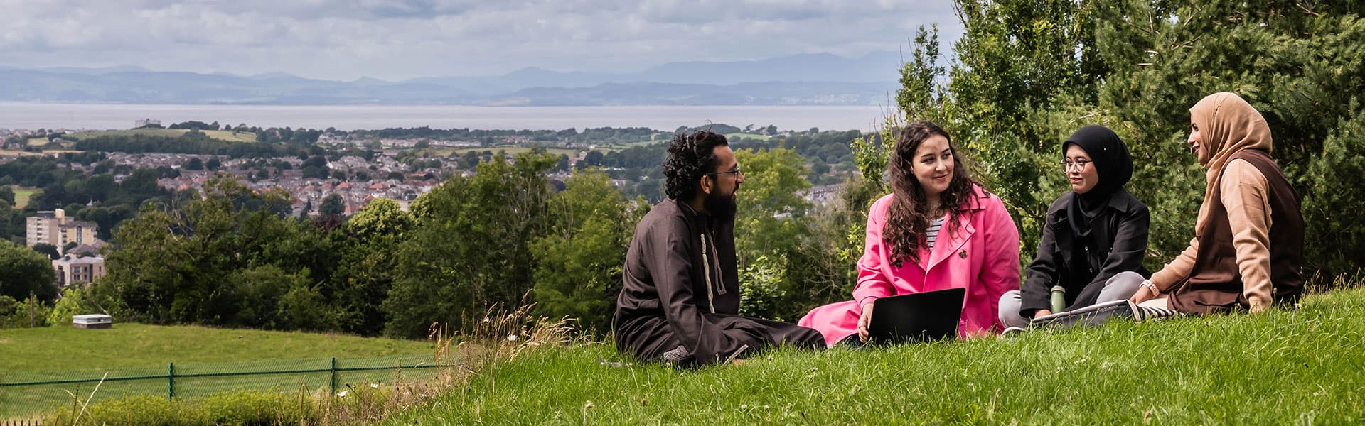 Staff sitting on grass, whilst surrounded by greenery. Morecambe Bay can be seen in the distance.