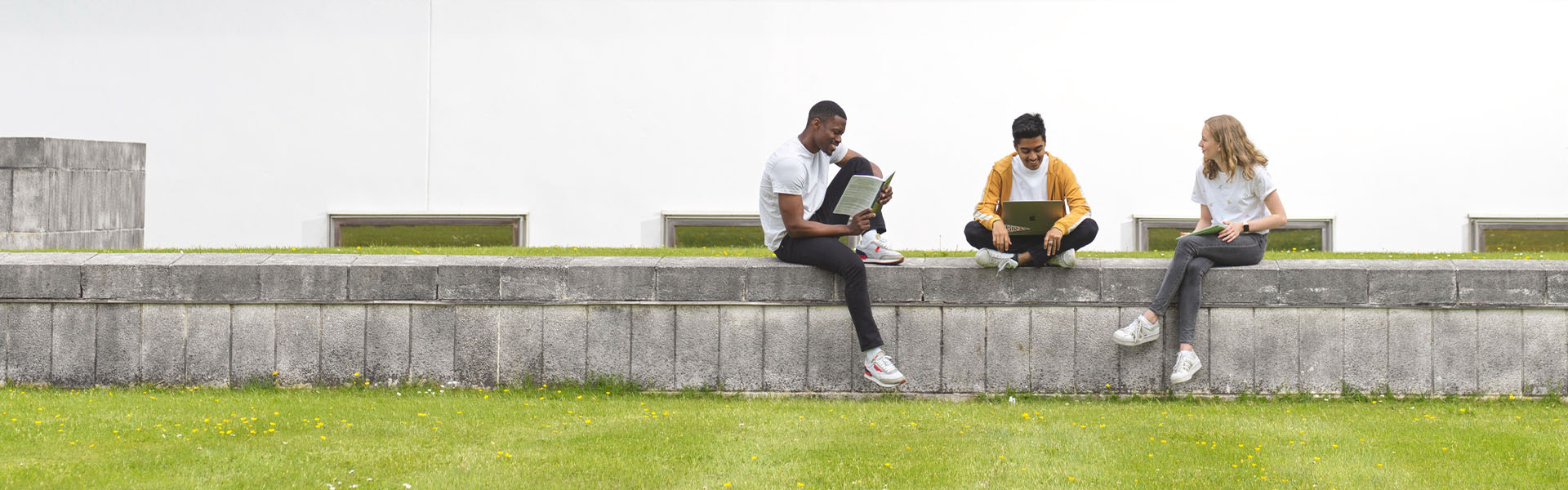 Three students sit on a wall, reading, near the Library.